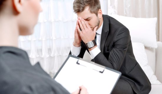 Young man with his psychologist Young man wearing a black suit sitting on a couch hiding his face with his hands, psychologist with clipboard listening to him and making notes during therapy session, selective focus