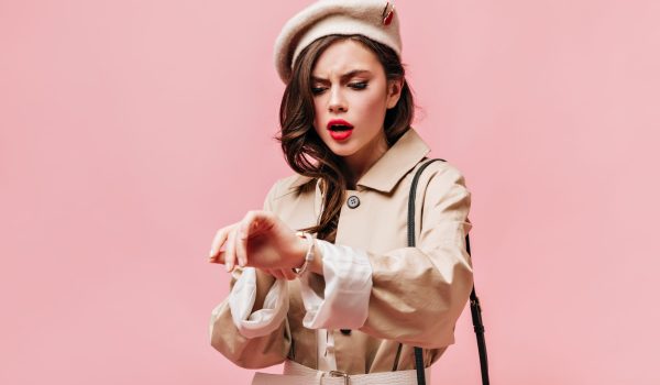 Brunette woman in beige trench coat and beret looks anxiously at wrist watch, posing on isolated background