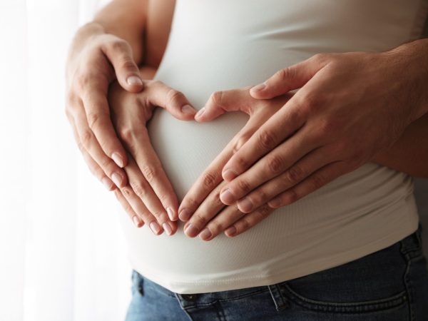 Close up portrait of man's and woman's hands making heart gesture over pregnant belly indoors